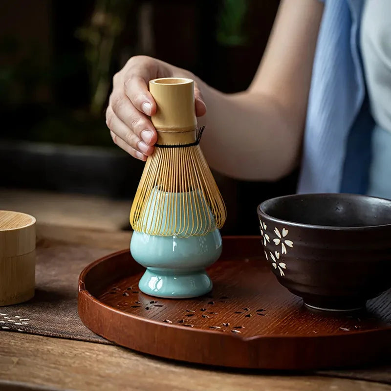 Person holding a bamboo matcha whisk above a bowl on a wooden tray, showcasing traditional tea preparation.