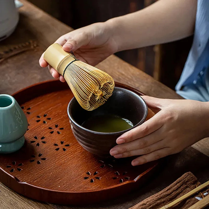 Close-up of a bamboo matcha whisk being used with a bowl of matcha tea on a wooden tray.