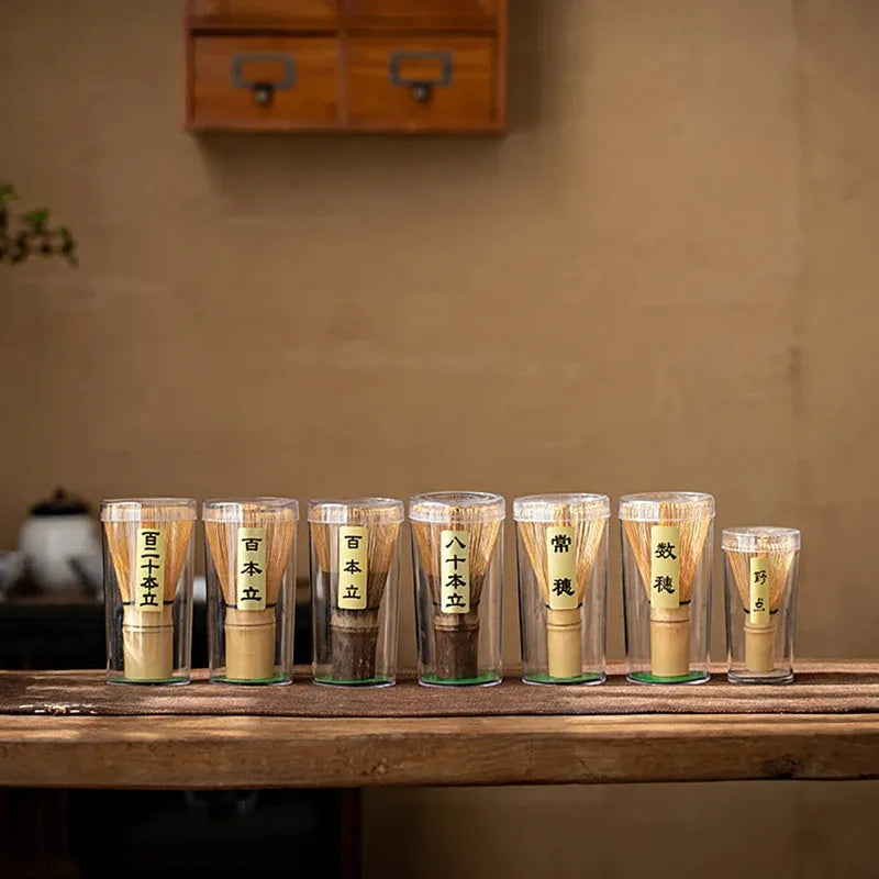 A collection of bamboo matcha whisks displayed in clear glass cups on a wooden table, showcasing traditional tea-making tools.